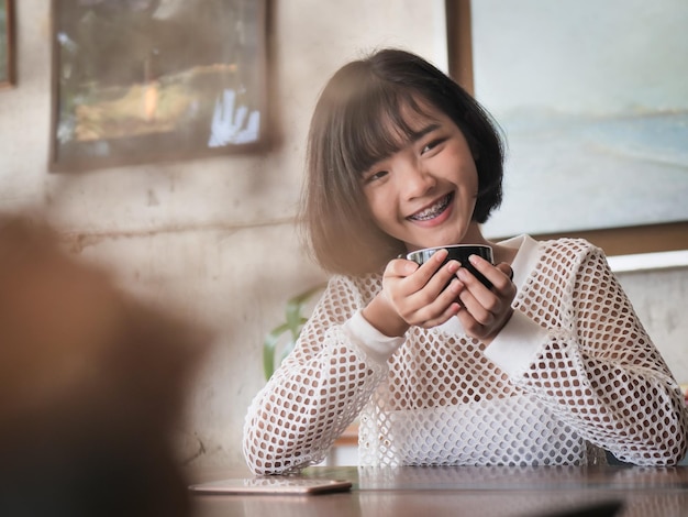 カフェに座っている間笑顔のティーンエイジャーの女の子がコーヒーカップを握っています