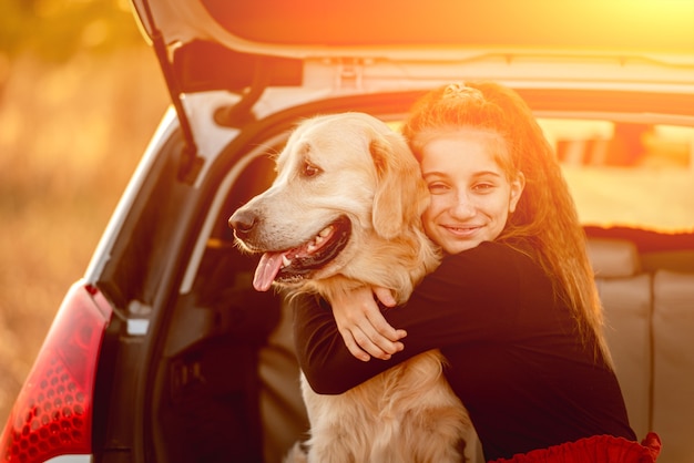 Smiling teenage girl hugging golden retriever dog in car trunk under sunlight