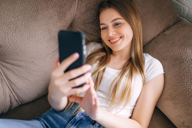 Smiling teenage girl relaxing while sitting on the couch at\
home using cell phone video chatting with friend