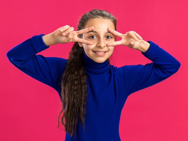 Smiling teenage girl  showing v-sign symbols near eyes isolated on pink wall