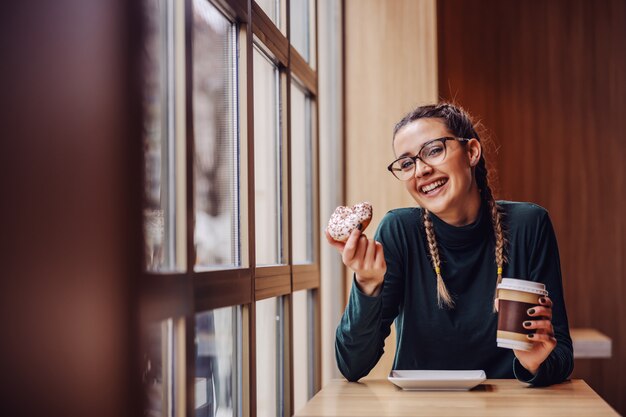 Smiling teenage girl sitting in cafe, holding doughnut and disposable cup with coffee on the break between classes.