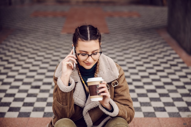 Улыбающаяся девочка-подросток сидит у парадного входа в старое здание, держит одноразовую чашку с кофе и разговаривает по телефону. занятия на выходные.