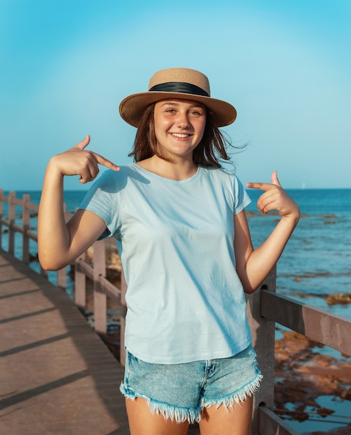 웃고 있는 10대 소녀는 밀짚 모자, 연한 파란색 티셔츠를 입고 그것을 가리키며 바다 옆 나무 보도에 머물고 있습니다. 티셔츠 모형