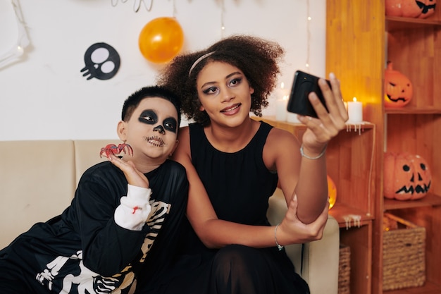 Smiling teenage girl taking selfie with her younger brother at Halloween party at home