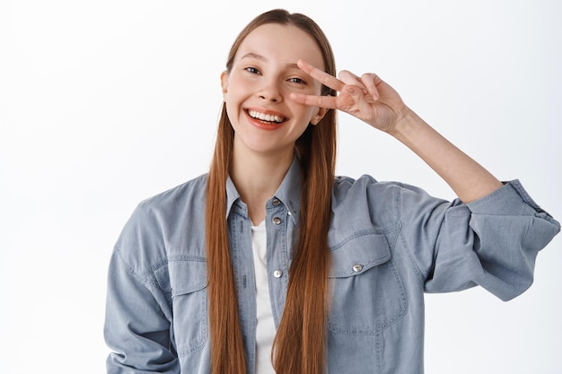 Adolescente sorridente con i capelli lunghi, mostra il gesto di pace con il segno a v e sembra felice, resta positivo, in piedi in abiti casual su sfondo bianco