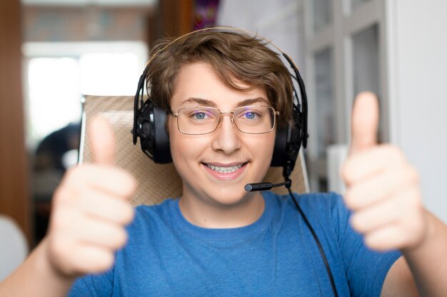 Smiling teenager boy in headphnes with microphone keeps thumbs up diring online video call.
