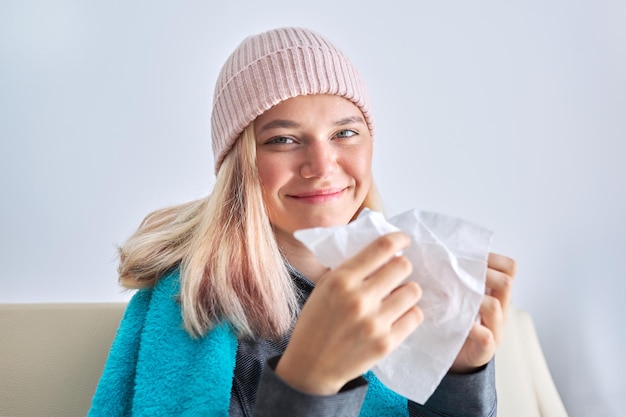 Smiling teenager girl sneezing in handkerchief female sitting at home covered with warm blanket and wearing knitted hat