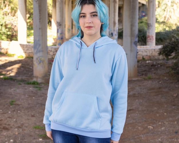 Smiling Teenager wearing light blue oversize hoodie and jeans stays under bridge  Blue haired teen girl stays outdoors against bridge pillars Clothing mockup