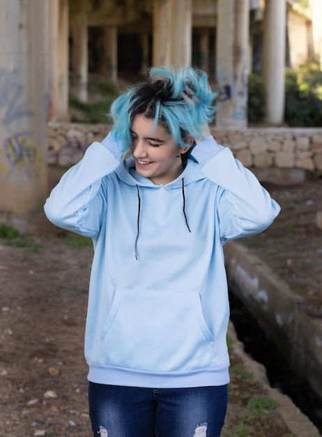 Smiling Teenager wearing light blue oversize hoodie and jeans stays under bridge Blue haired teen girl touch her hair outdoors against bridge pillars Clothing mockup