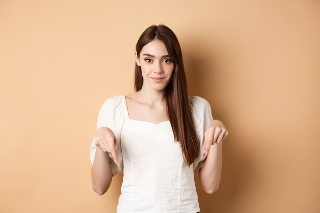 Smiling tender girl in dress pointing fingers down, showing advertisement on empty space, beige background.