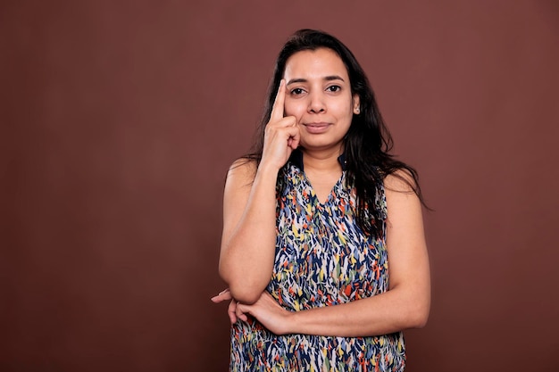 Smiling thoughtful indian woman posing portrait, model holding finger on face. Pensive person standing, looking at camera, front view studio medium shot on brown background