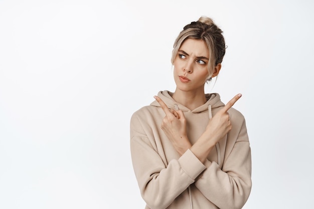 Smiling thoughtful woman pointing fingers sideways looking left with serious thinking face Girl making decision standing in beige hoodie against white background