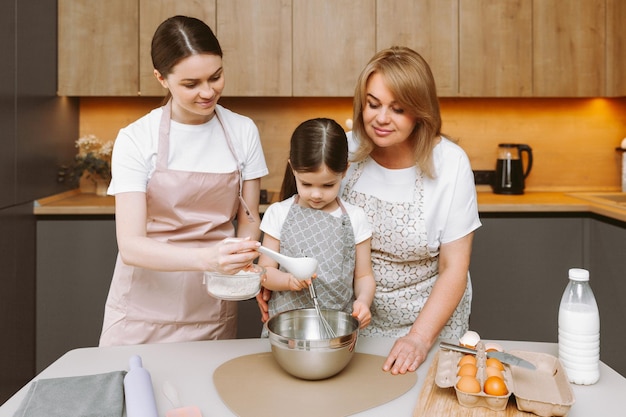 Smiling three generations of women have fun making dough in the kitchen A happy little girl with her mother and older grandmother are preparing pastries or cookies