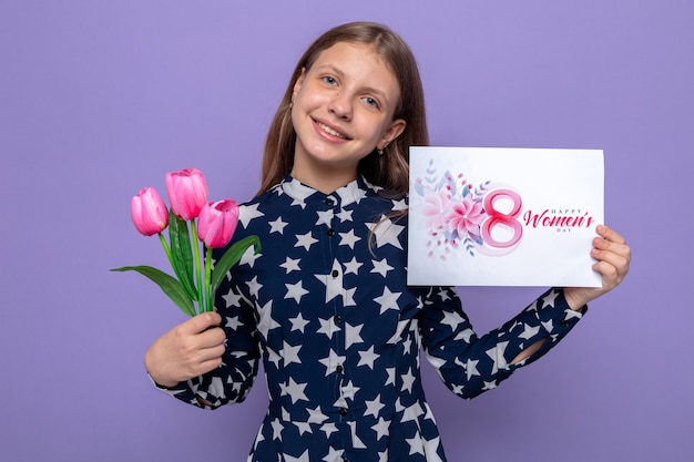 Smiling tilting head beautiful little girl on happy women's day holding flowers with postcard isolated on blue wall