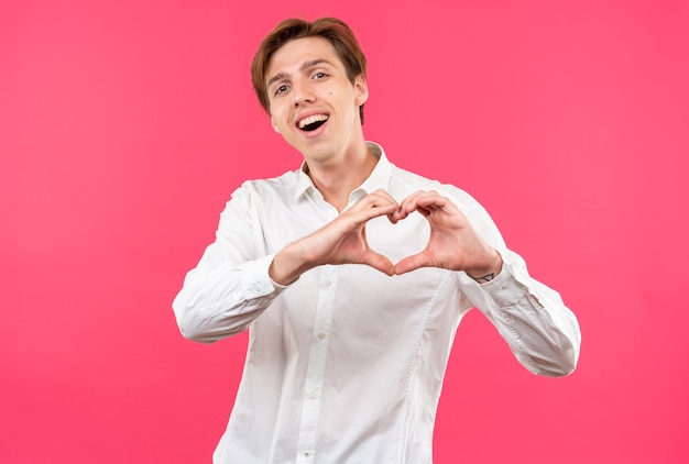 Smiling tilting head young handsome guy wearing white shirt showing heart gesture isolated on pink wall