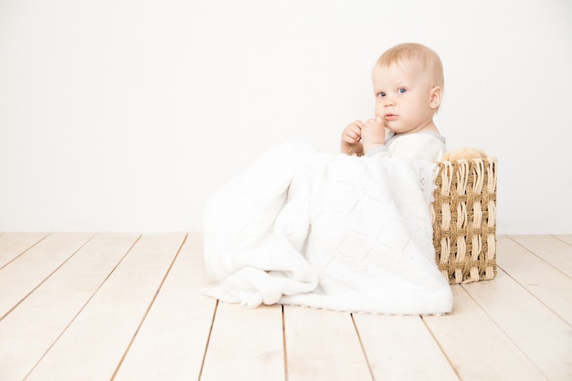 Улыбающийся малыш сидит в ведре с одеялом и игрушечным медведем, глядя в сторону на белом фоне.