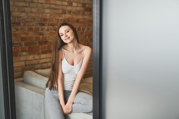 Smiling tranquil cute carefree young Caucasian woman sitting on the edge of the sofa indoors