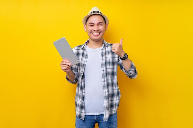 Smiling traveler tourist asian man in casual clothes hat holding tablet pc computer and showing thumbs up gesture on yellow background passenger traveling on weekends air flight journey concept