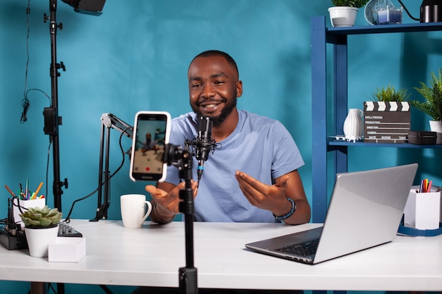 Smiling vlogger talking with audience in front of recording\
smartphone during online live show sitting at desk. content creator\
interacting with fanbase in studio looking at live video podcast\
setup.