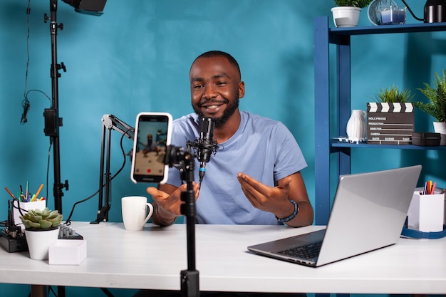 Smiling vlogger talking with audience in front of recording smartphone during online live show sitting at desk. Content creator interacting with fanbase in studio looking at live video podcast setup.