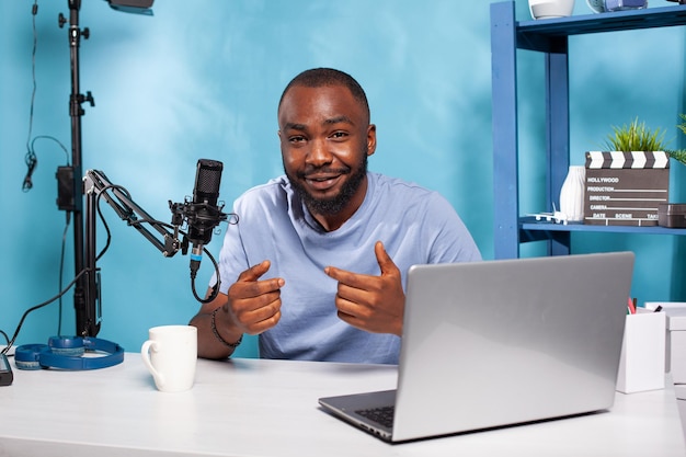 Smiling vlogger talking with audience during online live show sitting at desk with laptop and broadcasting setup. Content creator interacting with fanbase in recording studio with professional microph