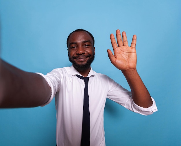 Smiling vlogger waving hello at camera in videocall conference on blue background. Wide angle pov of influencer taking a smartphone selfie doing hand gesture for social media.