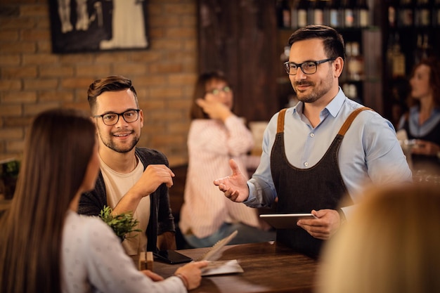 Smiling waiter with digital tablet taking order while talking with a guests in a bar