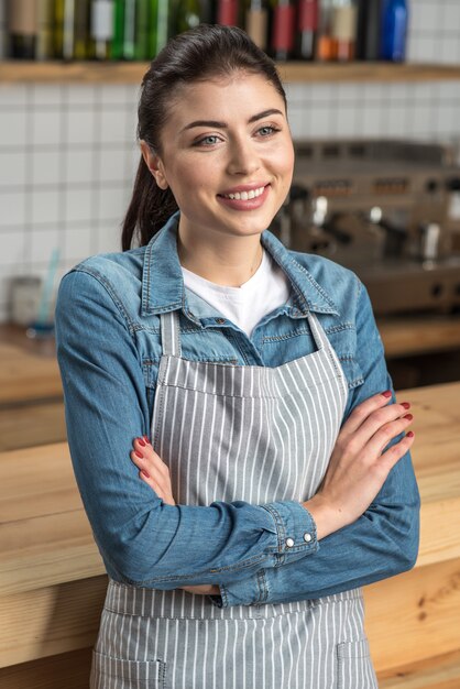 笑顔のウェイトレス。カフェの訪問者に戻る前に少し休憩しながら腕を組んでバーカウンターの隣に立っている経験豊富なプロの若いウェイトレス