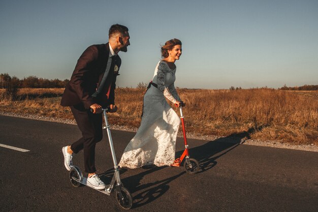 Smiling wedding couple riding a on scooters along the road outside the city at sunset.