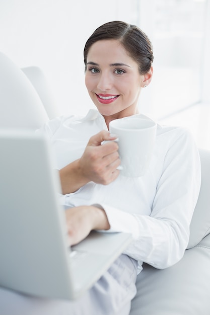 Улыбаясь хорошо одет женщина с ноутбуком и чашкой кофе на диване