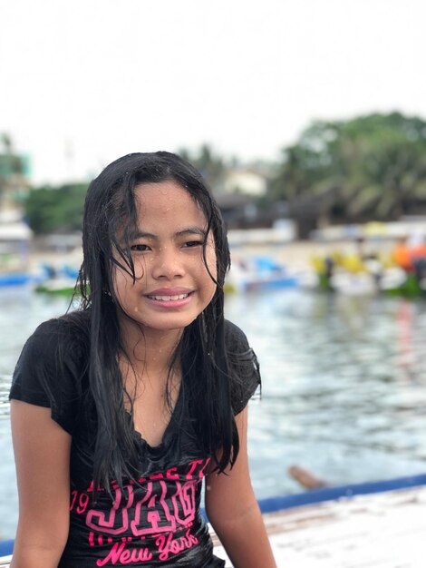 Photo smiling wet girl looking away while sitting at poolside against sky