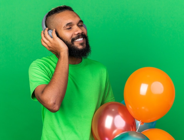 Sorridente con gli occhi chiusi giovane ragazzo afroamericano che indossa maglietta verde e cuffie in piedi dietro palloncini isolati sul muro verde