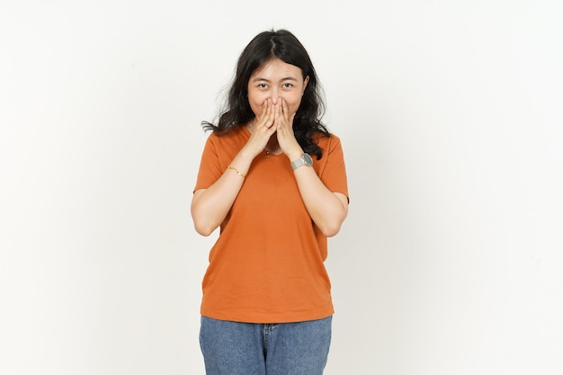 Sorridendo con la copertura della bocca di una bella donna asiatica che indossa una maglietta arancione isolata su bianco