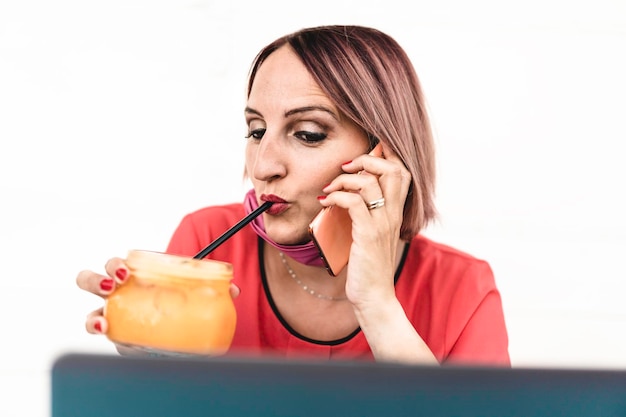 Foto donna sorridente di 40 anni che indossa una maschera protettiva aperta utilizzando laptop e telefono cellulare durante la fine dell'epidemia di coronavirus imprenditrice che lavora all'aperto mentre prepara una sana colazione