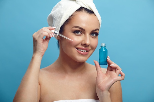 Smiling woman applies facial serum standing on a blue isolated background close up beauty portrait