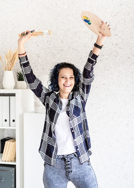 Smiling woman artist in her studio holding art palette and paintbrushes