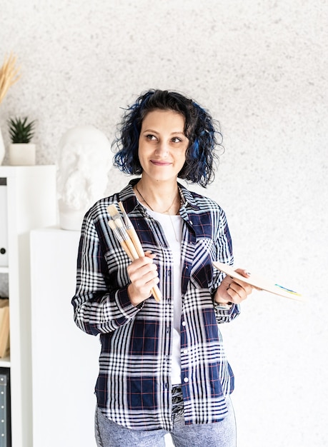 Улыбающаяся женщина-художник в своей студии держит художественную палитру и кисти