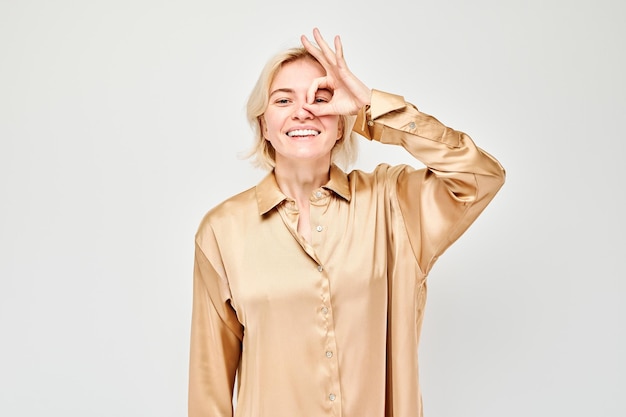 Smiling woman in beige blouse making OK gesture over eye isolated on light background