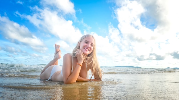 Donna sorridente in bikini che si trova sulla spiaggia tropicale, femmina felice nel colore bianco dello swimwear che gode delle vacanze estive che si trovano sulla sabbia bianca contro il cielo blu.