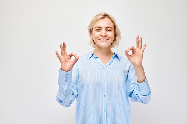 Улыбающаяся женщина в синей полосатой рубашке делает OK жест рукой на светлом фоне