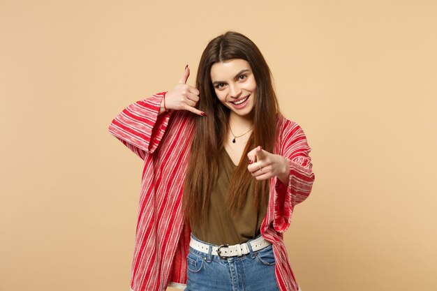 Smiling woman in casual clothes doing phone gesture like says call me back pointing index finger on camera isolated on beige background. People sincere emotions, lifestyle concept. Mock up copy space.