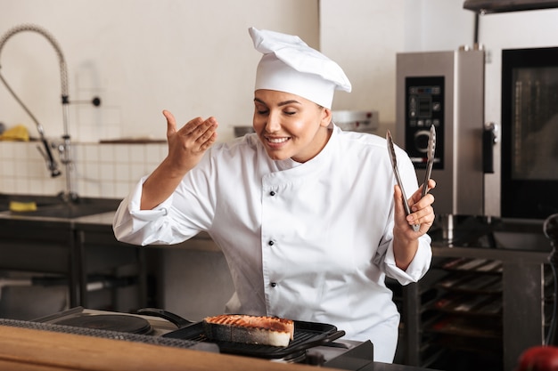 Smiling woman chef cook wearing uniform cooking delicious salmon steak standing at the kitchen