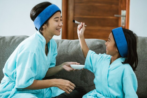 Smiling woman and child when using facial clay masks together while sitting on the sofa