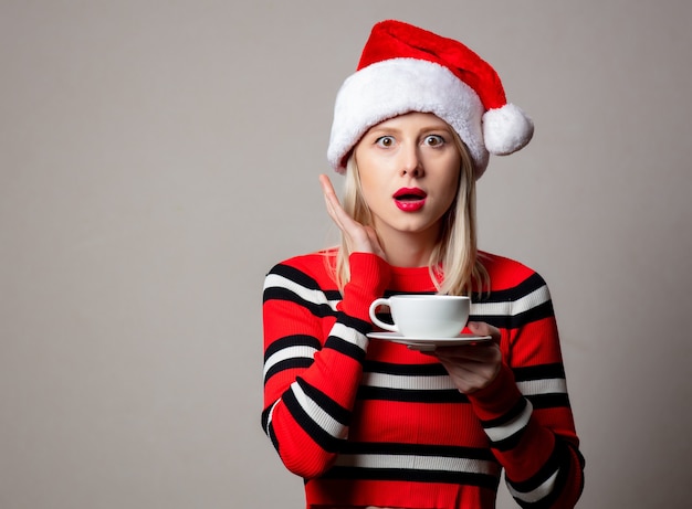 Smiling woman in Christmas hat with cup of coffee on grey wall