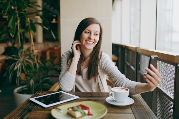 Smiling woman in coffee shop with cup of cappuccino, cake, doing selfie on mobile phone, relaxing in restaurant during free time. Female sitting with pc tablet computer rest in cafe. Lifestyle concept