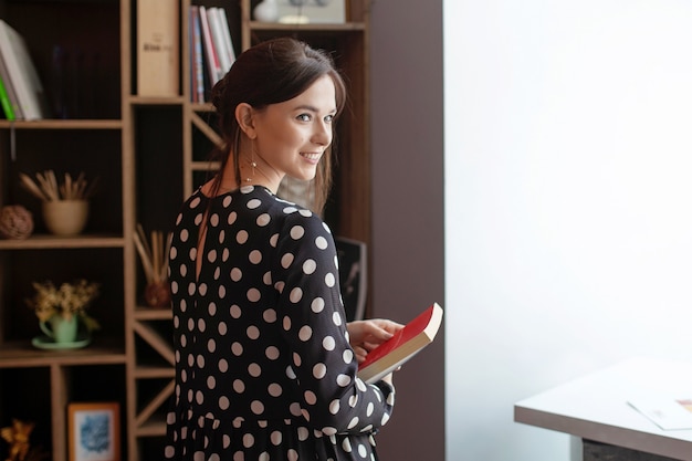 Smiling woman in dress standing in office. Businesswoman with book in hand.