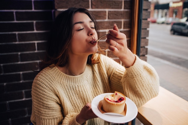 Photo smiling woman dressed warm sweater, enjoy eating dessert, tasty cupcake. snack food in street cafe