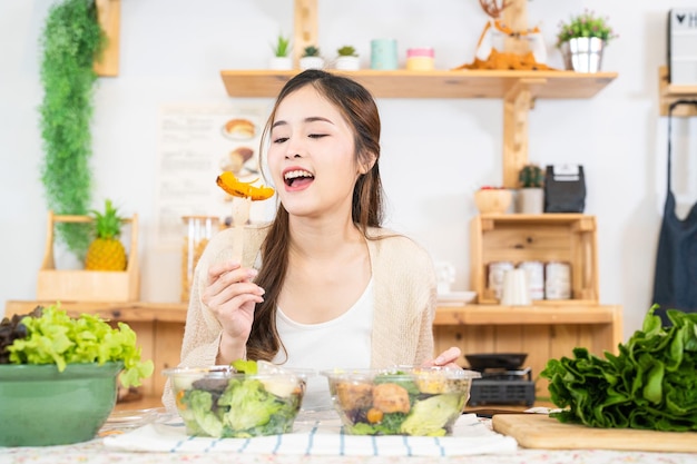Улыбающаяся женщина ест свежие здоровые салатные овощи Женщина сидит в кладовой на красивой внутренней кухне Чистая диетическая еда из местных продуктов и ингредиентов Свежий рынок