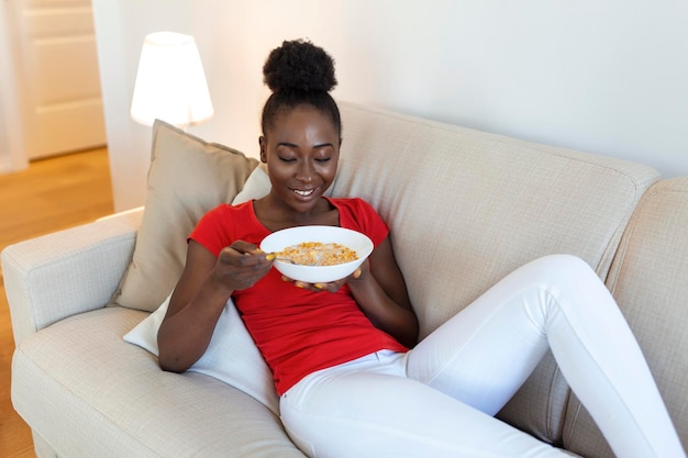 写真 リビングルームのソファでリラックスして朝の自宅で健康的な朝食を食べる笑顔の女性ソファに座ってボウルで食べ物を楽しんでいる美しい幸せなアフリカ系アメリカ人の女の子