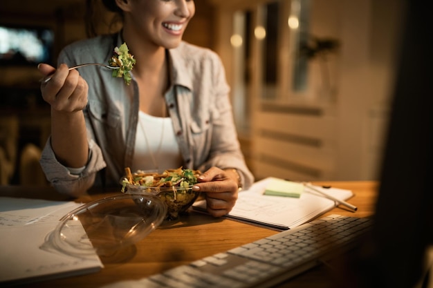 自宅のコンピューターで遅くまで働いている間、サラダを食べる笑顔の女性